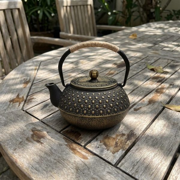 Théière en fonte 900 ml à picots arare rehaussés d'or, sur une table de jardin en teck, à l’ombre d’un feuillage